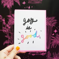 A hand holds up an LGBTQIA+ themed greeting card that says "Gay is Good" in a fun and colorful script.