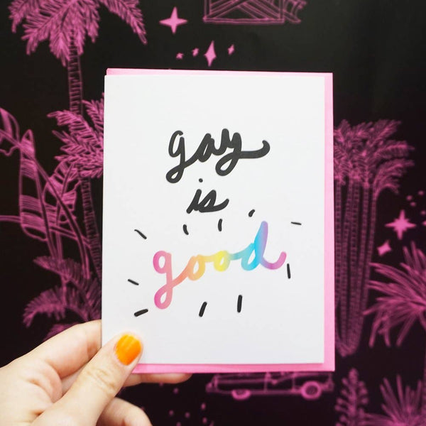 A hand holds up an LGBTQIA+ themed greeting card that says "Gay is Good" in a fun and colorful script.