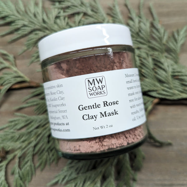 A clear glass jar containing rose colored clay sits on a wooden platform surrounded by cedar branches. The label features the MW Soapworks label and says "Gentle Rose Clay Net Wt 2 oz."