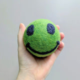 Hand holding a plush green wool dryer ball with a smiley face emoji felted design on it against a gray background