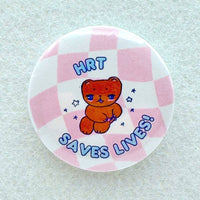 A pin features a cartoon bear that is surrounded by the text HRT SAVES LIVES on a pink checkered background.