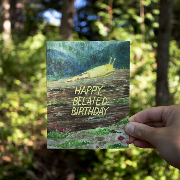 A hand holds up a greeting card against a PNW forest background. The card features a watercolor drawing of a banana slug crawling along the forest floor with the words "Happy Belated Birthday" across the middle.