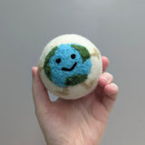 Hand holding a plush white wool dryer ball with a happy earth design against a gray background
