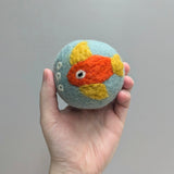 Hand holding a plush wool dryer ball with a happy goldfish design against a gray background