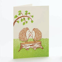 A white greeting card has two little hedgehogs facing each other on a tree stump. One has flowers in its paw to offer to the other.