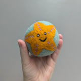 Hand holding a plush wool dryer ball with a felted happy starfish design against a gray background