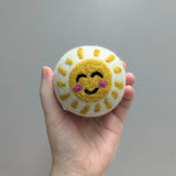 Hand holding a plush white wool dryer ball with a felted happy sun design against a gray background