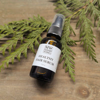 A 1 ounce glass bottle of MW Soapworks Healthy Hair Serum lays on a cedar branch on a wooden background.