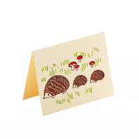 A greeting card shows three hedgehogs walking in a line in front of green grass and red mushrooms.