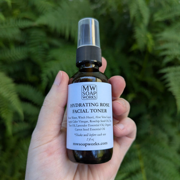 Hand holding a 2 ounce amber bottle of MW Soapworks Hydrating Rose Facial Toner against a green fern background.