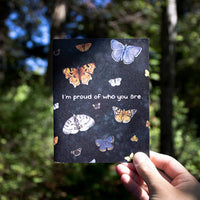 A hand holds a greeting card up against a pacific northwest forest. The card features watercolor illustrations of multiple moths and butterflies with the words "I'm proud of who you are" along the middle.