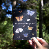 A hand holds a greeting card up against a pacific northwest forest. The card features watercolor illustrations of multiple moths and butterflies with the words "I'm proud of who you are" along the middle.