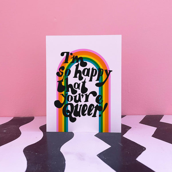 A greeting card sits against a pink and zigzag backdrop. The greeting card has a white background with a colorful rainbow across the card with big blocky funky black letters that say "I'm so happy that you're queer!"