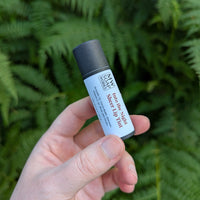 A hand holds up a sheer lip tint in front of PNW ferns. The lip tint is made by MW Soapworks and is a deeper red sheer color packaged in a biodegradable paper tube.