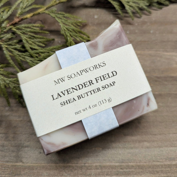 A purple and white swirled soap sits on a wooden background with cedar branches. The label indicates it is a four ounce Lavender Field Shea Butter Soap from MW Soapworks.