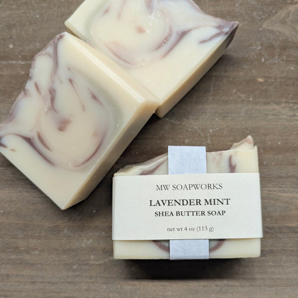 A 4 ounce bar of MW Soapworks Lavender Mint Shea Butter Soap sits on a wooden background. The soap is white with purple and green swirls from natural clay colorants.