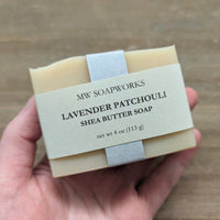 A hand holds up a 4 ounce bar of MW Soapworks Lavender Patchouli Shea Butter Soap against a natural wooden background.