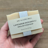 A hand holds up a 4 ounce bar of MW Soapworks Lavender Patchouli Shea Butter Soap against a natural wooden background.