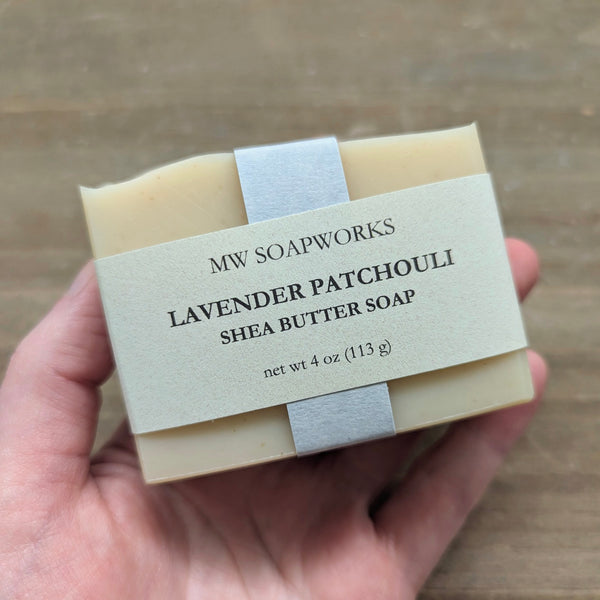 A hand holds up a 4 ounce bar of MW Soapworks Lavender Patchouli Shea Butter Soap against a natural wooden background.