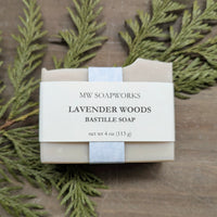 A four ounce bar of natural soap sits in front of a wooden background with cedar branches. The bar is light purple with a label that shows it is a Lavender Woods Bastille Soap bar from MW Soapworks.