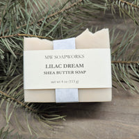 A light purple bar of MW Soapworks Lilac Dream Shea Butter Soap sits among branches from a Pacific Northwest forest.