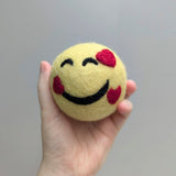 Hand holding a plush yellow wool dryer ball with a feeling loved emoji design felted on it against a gray background