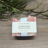 A bar of Lychee Fruit Shea Butter Soap sits on a wooden background with a cedar branch.