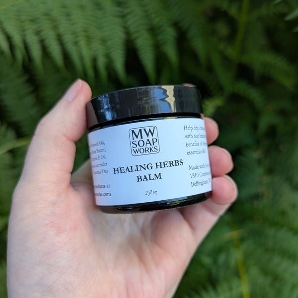 A hand holds a two ounce jar of Healing Herbs Balm from MW Soapworks against a green fern background.