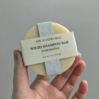 A hand holds up a round four ounce Solid Shampoo Bar from MW Soapworks. The bar is white and is four ounces of natural solid shampoo.