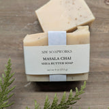 A bar of MW Soapworks Masala Chai Shea Butter Soap sits on a wooden platform. The soap has a creamy off white base color with real ground tea leaves mixed in.