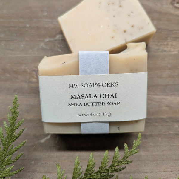 A bar of MW Soapworks Masala Chai Shea Butter Soap sits on a wooden platform. The soap has a creamy off white base color with real ground tea leaves mixed in.