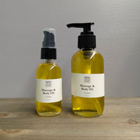 Two clear glass bottles of MW Soapworks Massage and Body Oil sit on a wooden shelf. The bottles contain 2 ounces and 4 ounces of Cosmos Massage and Body Oil.