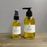 Two clear glass bottles of MW Soapworks Massage and Body Oil sit on a wooden shelf. The bottles contain 2 ounces and 4 ounces of Cosmos Massage and Body Oil.