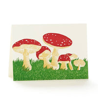 A letterpress greeting card features numerous red cap mushrooms in green grass.