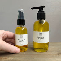 Two clear glass bottles of MW Soapworks Massage and Body Oil sit on a wooden shelf. The bottles contain 2 ounces and 4 ounces of San Fran Massage and Body Oil and have pump lids.