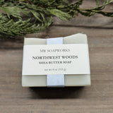 A 4 ounce bar of MW Soapworks Northwest Woods Shea Butter Soap sits near a cedar branch against a wooden background.