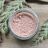 A clear glass jar of rose colored clay is open on a wooden background surrounded by cedar branches. 