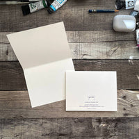 An open greeting card sits open while another shows the back. The inside is blank and the back shows the artist Yardia's logo and information.