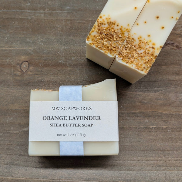 Bars of MW Soapworks Orange Lavender Shea Butter Soap sit on a natural wood background. In the corner you can see the tops of two bars, showing sprinkled ground citrus peel along the top of the bar soap.