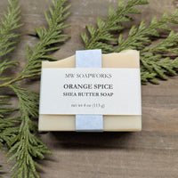 A 4 ounce bar of natural soap sits on a cedar branch. The label shows it is a bar of Orange Spice Shea Butter Soap from MW Soapworks.