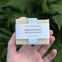 A hand holds a four ounce bar of Orange Spice Shea Butter Soap from MW Soapworks in front of a fern background.