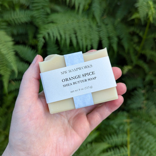 A hand holds a 4 ounce bar of Orange Spice Shea Butter Soap from MW Soapworks in front of a Pacific Northwest fern background.