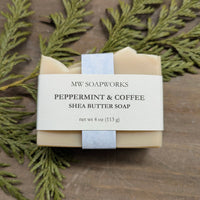 A tan bar of soap sits on a natural background of wood and cedar branches. The label shows it is a four ounce Peppermint and Coffee Shea Butter Soap from MW Soapworks.