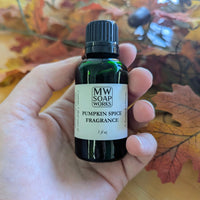 A hand holds up a green one ounce bottle of Pumpkin Spice Fragrance from MW Soapworks in front of colorful fall leaves.