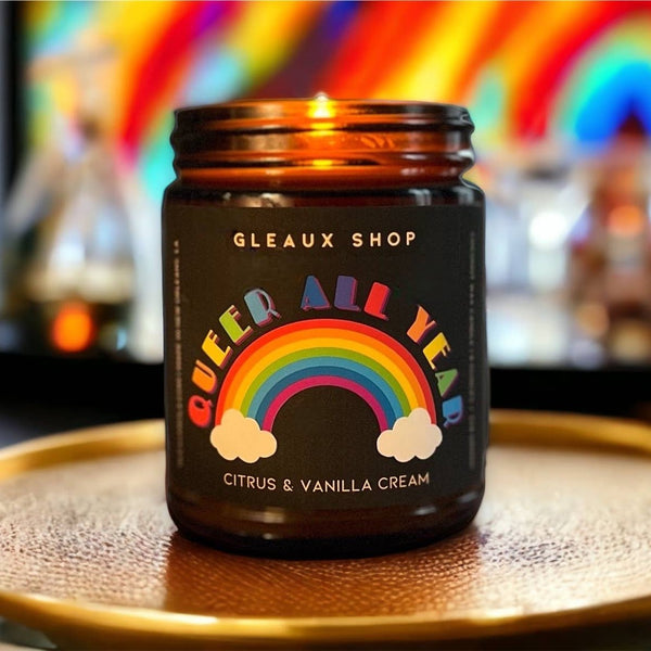 A candle packaged in an amber jar sits on a coaster on a table. The label reads "Queer All Year" with a fun rainbow design.