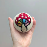 Hand holding a plush white wool dryer ball with a rainbow colored mushroom design against a gray background