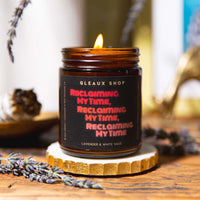 A candle packaged in an amber jar sits on a coaster on a table. The label reads "Reclaiming my time, Reclaiming my time, Reclaiming my time."