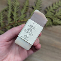 A side view of the Lavender Field Natural Soap from MW Soapworks. The soap is white and purple swirls and has a label with logos that indicate it is made in Washington state and that the company is Queer and Disabled owned and that they give back.