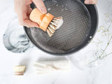 Person holding a wooden brush over a frying pan on a light background