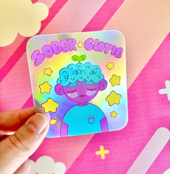A sticker features a cartoon queer cutie surrounded by stars and the words "Sober Cutie" above them in bubble letters.